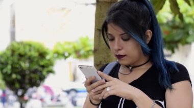 Gülümseyen güzel bir genç kadın. Mavi saçlı üniversiteli kız parkta sosyal medyasına bakıyor, bir ağacın yanında. Parkta cep telefonuna bakan bir kız. Teknoloji, iletişim ve eğitim kavramı