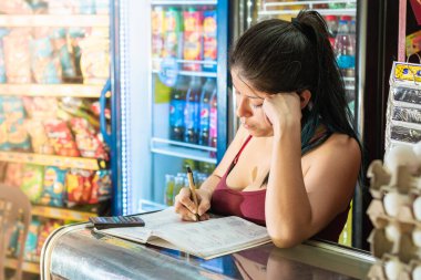Latin bir kadın marketinin hesaplarını deftere yazarken çok düşünceli davranmış. Cam vitrine yaslanmış, satışları düşünen bir kız. Ekonomi ve iş konsepti.