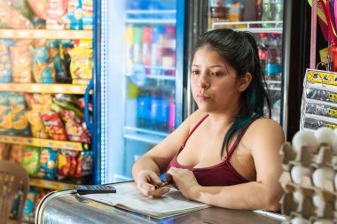 Latin bir kadın marketinin hesaplarını deftere yazarken dikkati dağılmış. Cam vitrine yaslanmış, satışları düşünen bir kız. Ekonomi ve iş konsepti