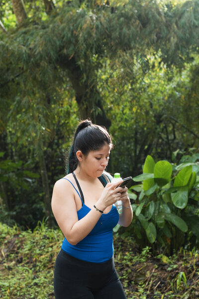 beautiful latin girl walking on a trail in the middle of a forest in colombia. sending messages with her cell phone and taking pictures. influencer walking through the jungle without enjoying nature.