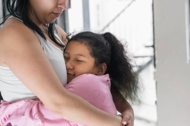 Yüzü annesinin göğsüne bastırılmış güzel kahverengi tenli Latin kız gülümseyerek gözlerini kapatırken kalbini dinliyor ve mutlu bir jest yapıyor. Küçük kız annesine sarılıyor..