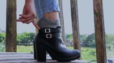 Bir kızın ayaklarının, tahta parmaklıklı bir balkonda ayak bileği botlarını giydiği yakın plan bir detay. Kot pantolon ve sentetik deri ayakkabılarla yürüyüşe çıkmak için ayakkabılarını giyen genç bir kadın. moda konsepti
