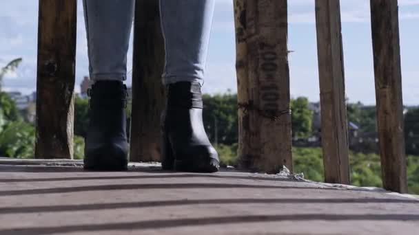 一个穿着脚踝靴 站在有木制栏杆的阳台上的女孩的脚部的特写 穿着蓝色牛仔裤和人造皮鞋散步的女人 时尚概念 — 图库视频影像