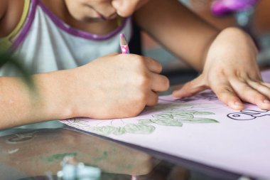 Kahverengi tenli, düşük gelirli Latin bir kızın pembe bir posterde renkli kalemlerini boyamasının detaylı görüntüsü. Kız çalışmalara ve çizimlere konsantre oluyor. yoksul insan kavramı.