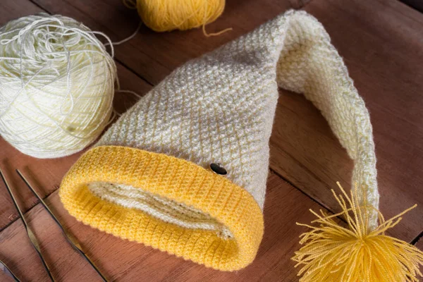 still life of a yellow crocheted hat with materials for designing and knitting garments, placed on a wooden background. rolls of wool and steel needles for home knitting.