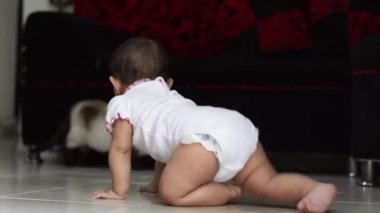 Kahverengi tenli güzel Latin bir kız bebek, oturma odasında yerde sürünmeyi öğreniyor, sekiz aylıkken, arka planda bir kedi ve kırmızı bir kanepe ile. Kız ilk adımlarını atıyor