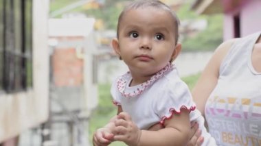Kahverengi tenli güzel Latin kız, çok mutlu, annesi tarafından taşınırken ellerini sallıyor ve mutlu jestler yapıyor. Kadın bebeğini sokakta gezdirir..