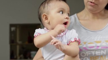 Beautiful latina baby girl with brown skin, on her mother's arms looking very surprised towards the street. eight months old girl with a white romper suit looking scared at the streets.