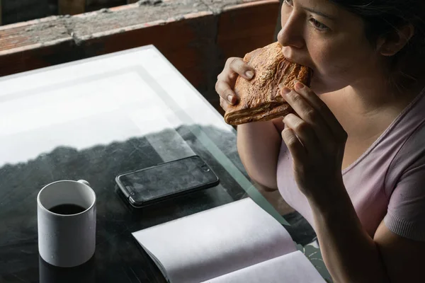 beautiful latina woman eating a sandwich for breakfast before going to college, drinking coffee in a white mug and holding her cell phone next to a blank book ready to copy text. office concept