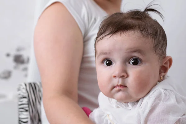 beautiful baby with big eyes, sitting on her mother\'s lap after receiving food. little girl looking curiously with her mother behind her. baby dressed in white