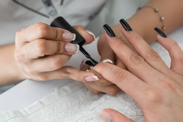 close-up detail shot of a manicurist\'s hands painting the nails with a black nail polish. designing and beautifying the nails of a woman in the beauty salon. varnishing the nails. dark style.