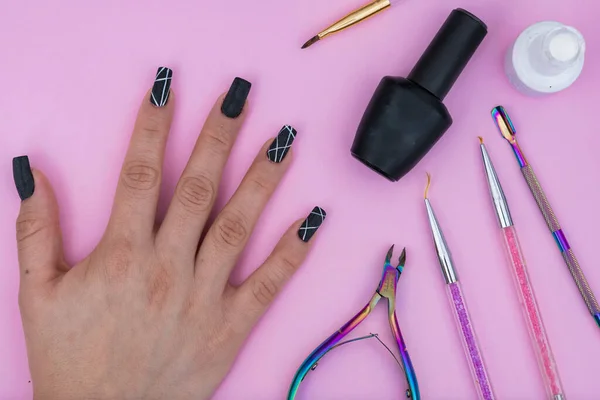 hand of a Latin girl, surrounded by basic elements for a manicure, placed on a pink background. black and white nail polish, colored brushes, cuticle pusher and cuticle nippers.