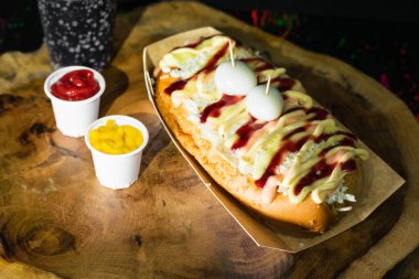 top view of a hot dog with sauces, cheese and two quail eggs on top served on a wooden tray and next to it are two plastic cups with ketchup and mustard, next to the black soda.