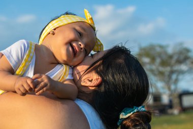 close-up of a beautiful latin baby girl being held by her mother, nodding her head, smiling and holding her cheeks together, giving affection to each other. beautiful summer day with sky background.