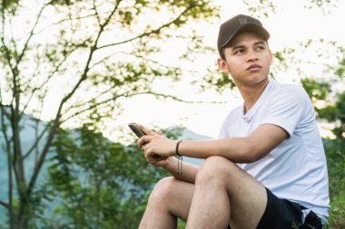 Genç Latin bir adam, bir dağın tepesinde oturuyor, cep telefonu elinde ve arka planda güzel altın bir gündoğumu, mutlu bir şekilde çevresindeki doğaya bakıyor..