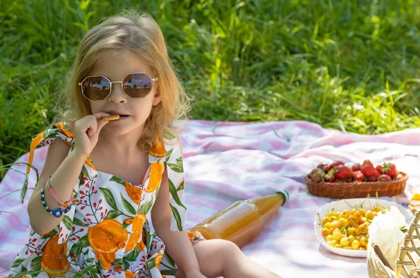 A happy 5-year-old girl is sitting on a mat and enjoying cookies at a picnic in the park. Enjoying outdoor recreation. Child in sunglasses
