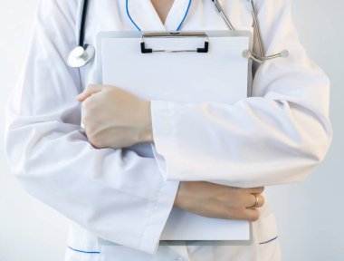 A medical worker in a white coat with a stethoscope holds a clipboard in his hands