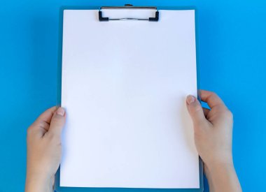 Clipboard with blank white sheet on blue background. View from above. Female hands holding clipboard, place for text