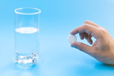 A male hand holds a pill near a glass of water on a blue background