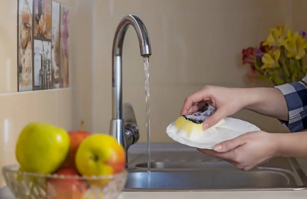 Womens hands wash a white plate with a sponge under running water in the kitchen sink. Washing dishes at home