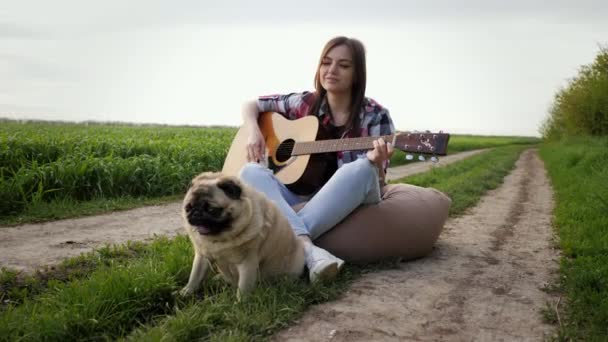 Mujer cantando y tocando la guitarra con perro pug sentado en la silla bolsa en el campo verde — Vídeos de Stock