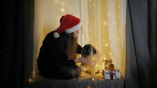 Liten söt tonåring flicka i Santa hatt leka med sin roliga mops hund, sitter på fönsterbrädan dekorerad med en krans — Stockvideo