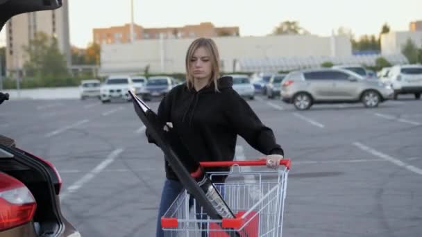 Vrouw draagt een moderne grote tv in een supermarkt trolley en probeert om het te laden in de kofferbak van een auto op de parkeerplaats — Stockvideo