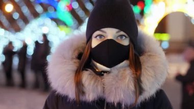 Koruyucu maskeli genç bir kadının portresi Noel ışıklarının ve çelenklerin arka planında duruyor ve kameraya bakıyor.