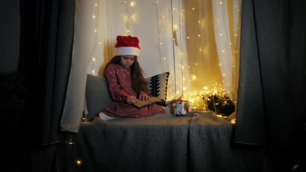 Kleines Mädchen schreibt einen Brief an den Weihnachtsmann mit Wünschen, die auf der mit Girlanden geschmückten Fensterbank sitzen — Stockvideo