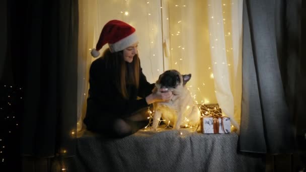 Gadis remaja bertopi santa bermain dengan anjing pug lucu, duduk di jendela dihiasi dengan karangan bunga — Stok Video