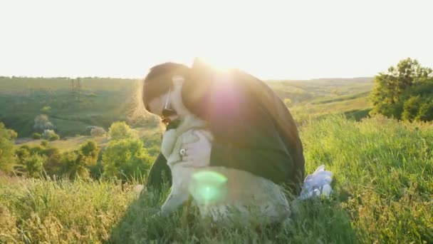 Właścicielka przytula się i całuje psa o zachodzie słońca. Pug i kobieta siedzą w przyrodzie i odpoczywają na wzgórzach, kochają zwierzęta — Wideo stockowe