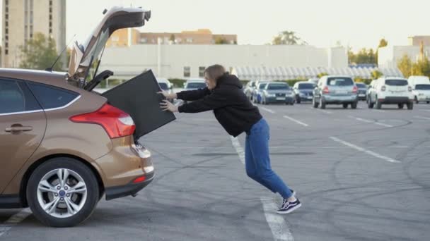 Vrouw grappig proberen om aangekochte moderne tv in auto kofferbak bij supermarkt parkeren — Stockvideo