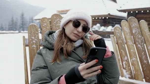 Fashionable young woman in sunglasses use mobile phone under a snowfall in the mountains at a resort — 图库视频影像
