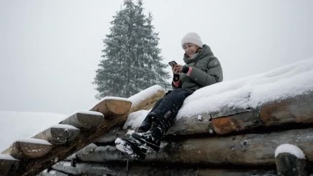 Young woman use mobile phone under a snowfall in the mountains at a resort, sitting on the snow. — Stockvideo