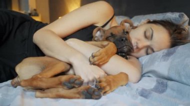 Yorgun genç bir kadın köpeğiyle birlikte evde uyuyor, sevimli köpek yavrusunu yakından kucaklıyor.