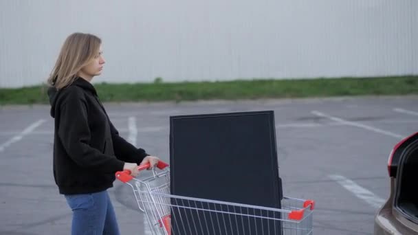 Vrouw draagt een moderne grote tv in een supermarkt trolley en probeert om het te laden in de kofferbak van een auto op de parkeerplaats — Stockvideo