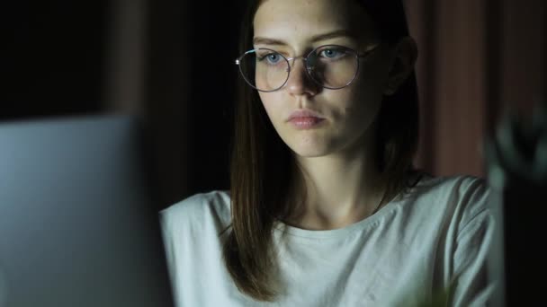 Ένα έφηβο κορίτσι με γυαλιά κοιτάζει την οθόνη, παίζει ένα παιχνίδι, διαβάζει, μελετάει αργά το βράδυ. — Αρχείο Βίντεο