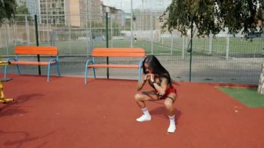 Atletik kadın çömeliyor, yazın spor sahasında spor bandıyla bacaklarını güçlendiriyor.