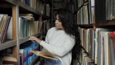 Genç bir kadın eski kütüphanedeki raftan bir kitap seçiyor, kitaplığa bakıyor.