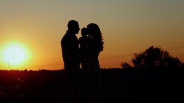 Ebeveynler bebeklerini öper ve kucaklarlar. Gün batımında tanınmaz bir aile silueti. Aile değerleri.