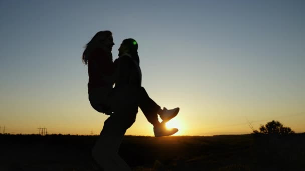 Молодая девушка прыгает на своего парня и они кружат на фоне заката, силуэт счастливой пары — стоковое видео