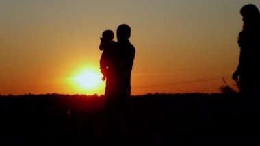Bakıcı baba bebeği kollarında tutar, anne gelir ve aileyi kucaklar, gün batımında tanınmaz bir siluet, baba sevgisi