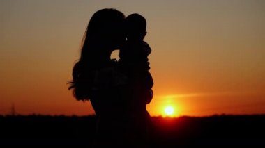 Şefkatli anne bebeği kollarına alır ve onu öper. Gün batımında tanınmaz bir siluet, anne sevgisi.