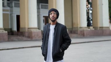 Gözlüklü ve şapkalı yakışıklı bir Arap caddede kameraya ağır çekimde bakıyor.