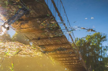 Nehrin üzerindeki ahşap kablo köprüsünde aşağıdan bir broş ve güneşli bir günde su yüzeyinde bir köprü yansıması var.