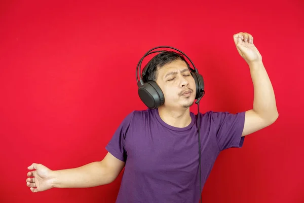 Asyalı bir adam kulaklık kullanarak şarkı dinlerken eğleniyor ve dans ediyor. kırmızı arkaplan. 