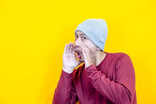 Asyalı bir adam bereli ve kırmızı tişörtlü, fısıldayan bir ifadeyle. Sarı arkaplan.