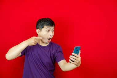 Asyalı yakışıklı, mor tişörtlü, kızgın yüzlü ve akıllı telefonunu işaret eden bir adam. kırmızı arkaplan.