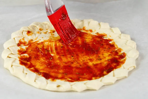 Proceso de elaboración de pizza. Vista superior de la base de masa de pizza con una corteza de pastel trenzado. cepillado con pasta de tomate —  Fotos de Stock