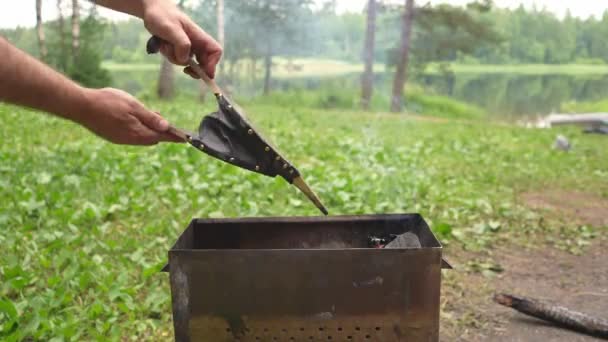 Un hombre sopla un fuego con el fuelle de chimenea de madera dura en la barbacoa. Descanse en el bosque en un viaje de pesca junto al lago. — Vídeos de Stock